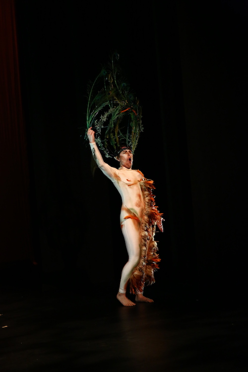 Jules Skloot wears a unitard with feathers trailing down the midline to the foot. A headpiece made of grass and plants sits atop his head.